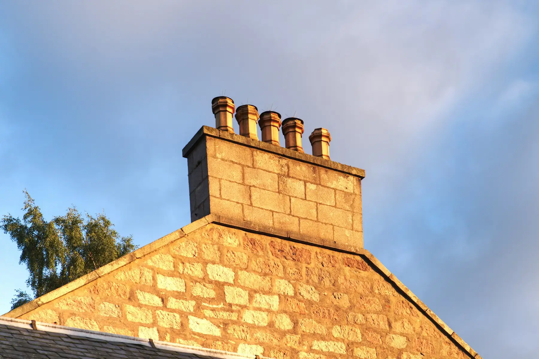 Stone chimney stack with five clay pots above a slate roof, lit by low sun