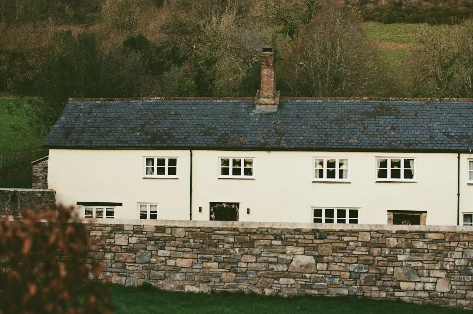 A long white farmhouse with a slate roof behind a dry stone wall