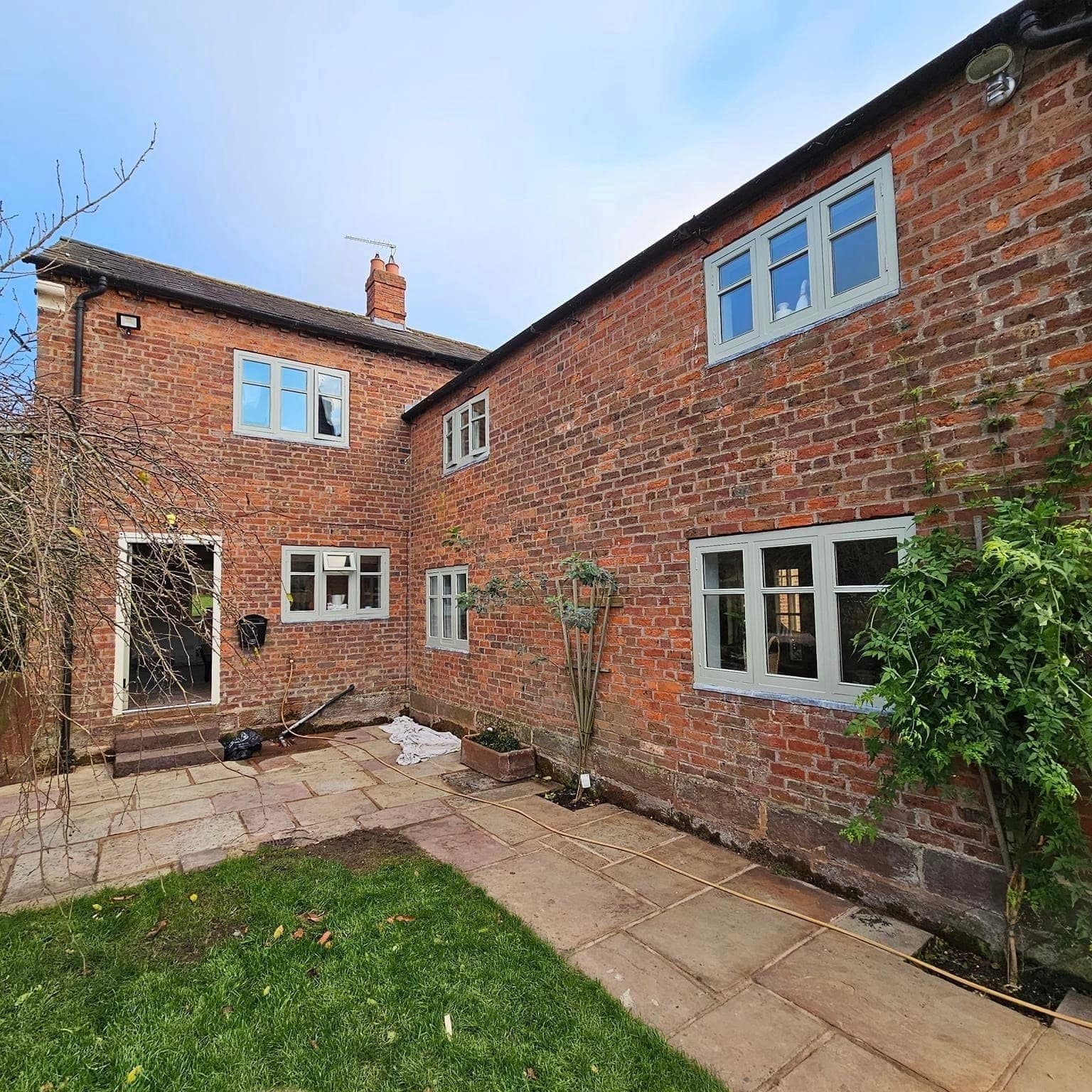 The courtyard side of a red-brick farmhouse with pale grey window frames and a paved yard