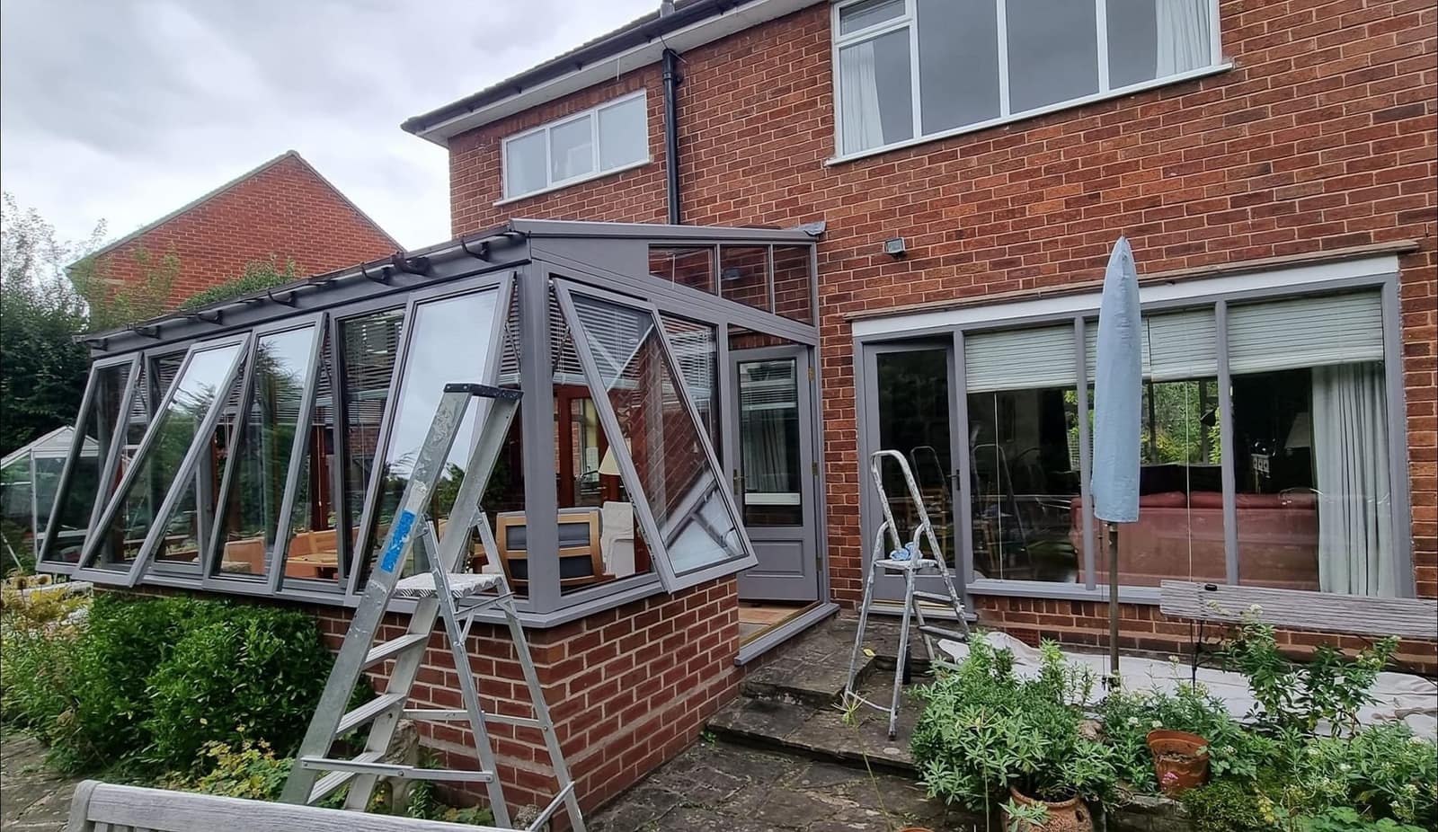 The back of a red-brick house with a grey-framed conservatory and stepladders set up in the garden