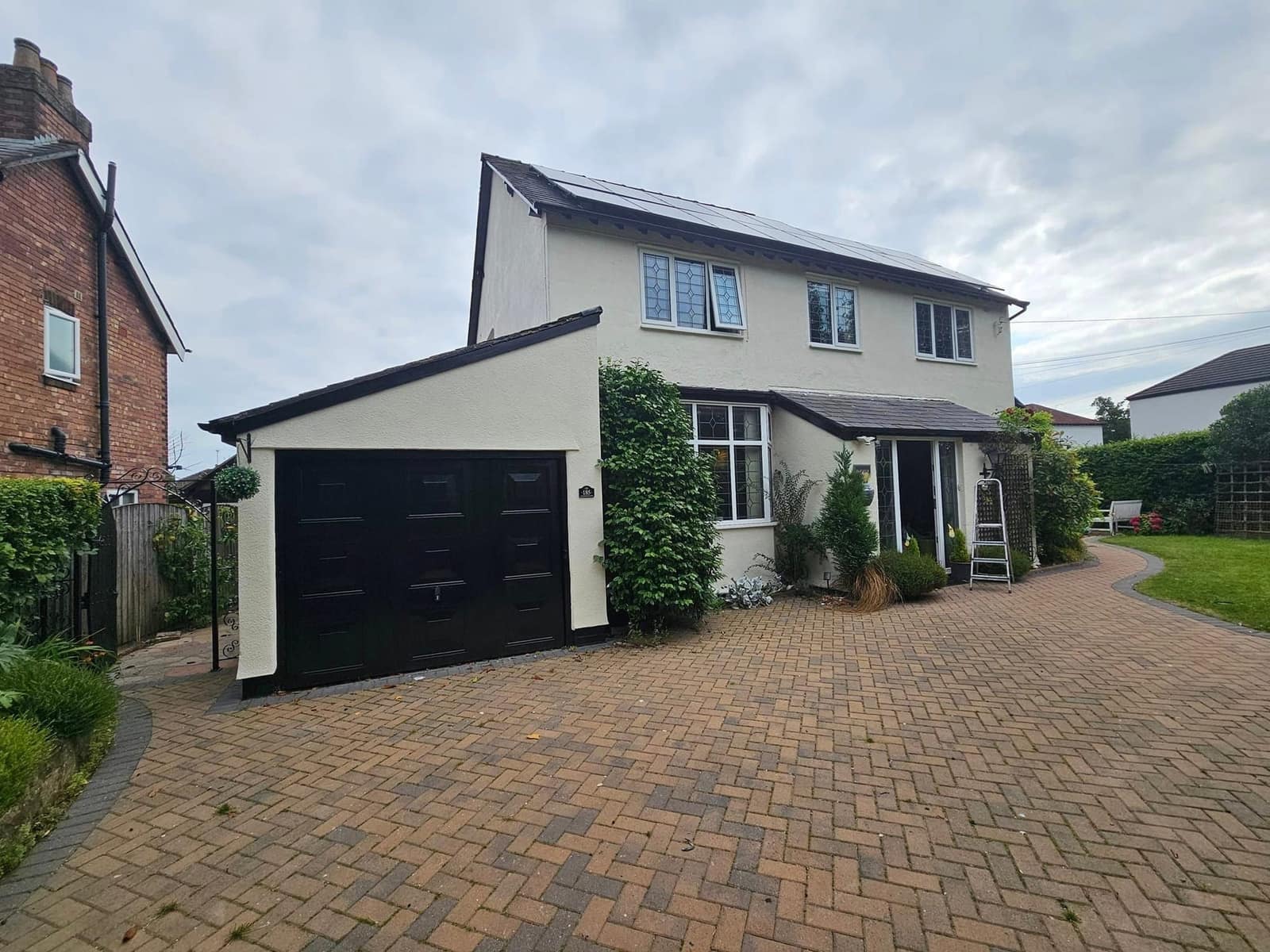 A detached house finished in cream render with a black up-and-over garage door, seen from the block-paved drive
