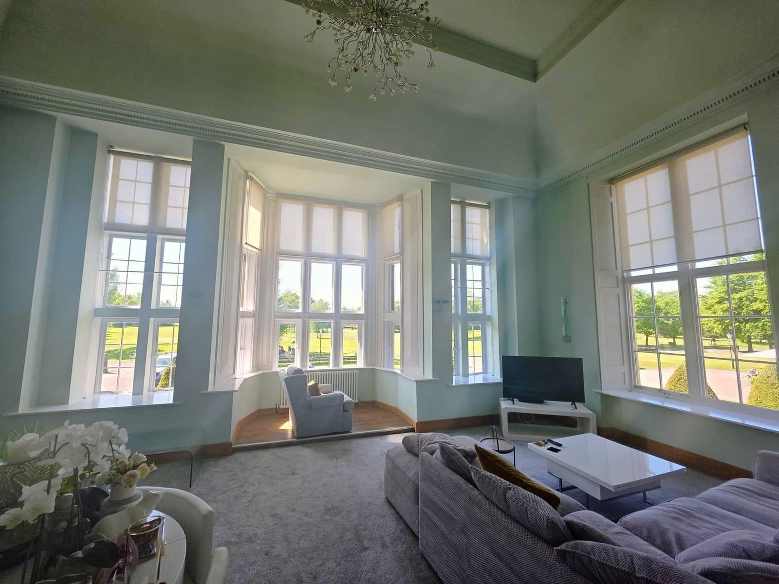 A large lounge in pale sage green with a deep bay window, a panelled ceiling and a crystal chandelier