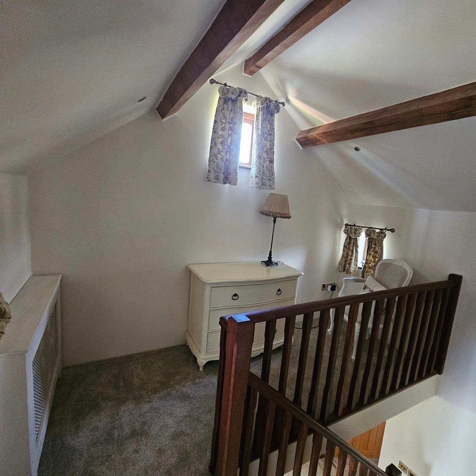 A cottage bedroom under dark exposed beams, with a dark stained balustrade at the top of the stairs