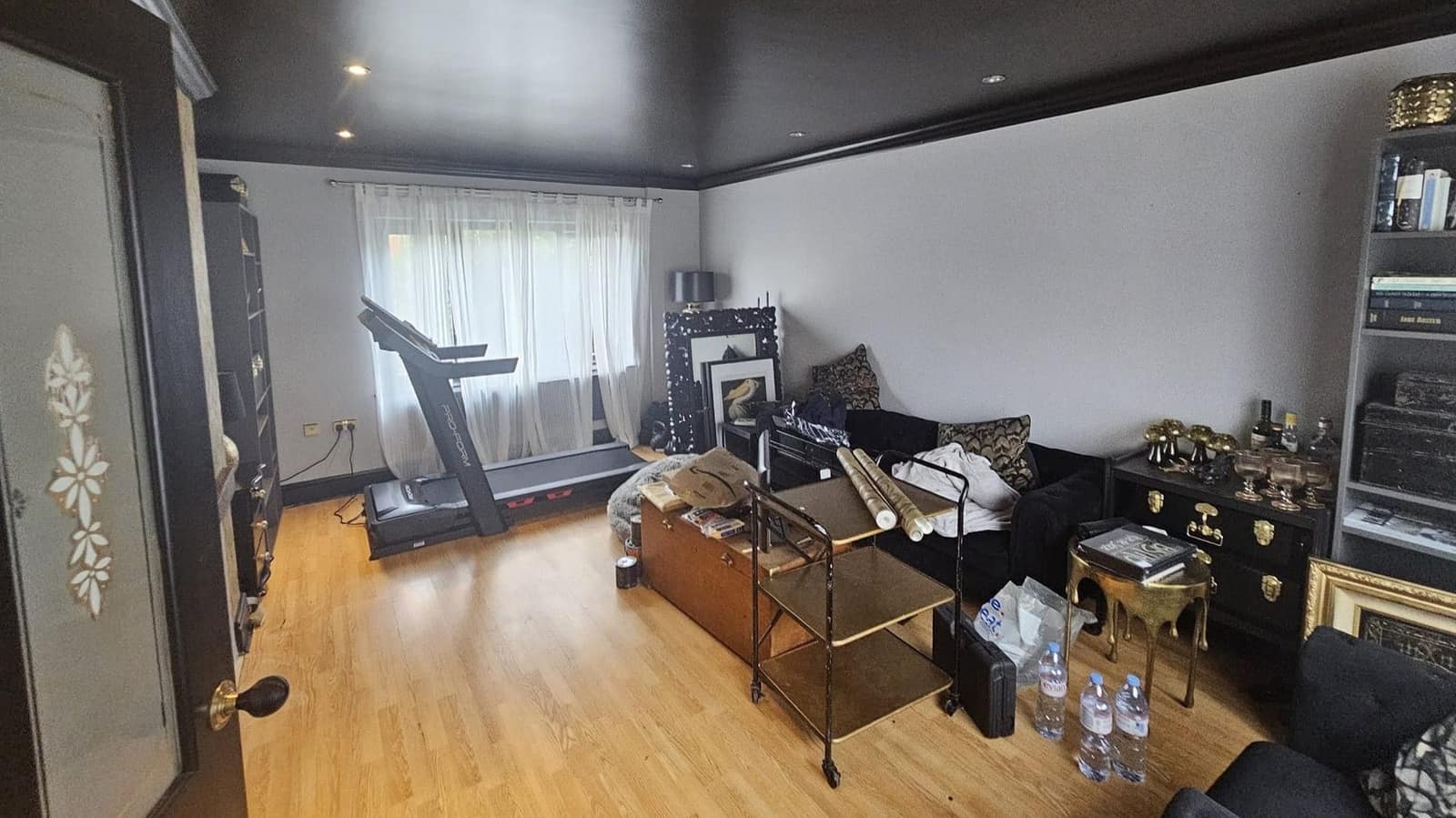 The same room from the door: black ceiling, pale walls, a treadmill by the window