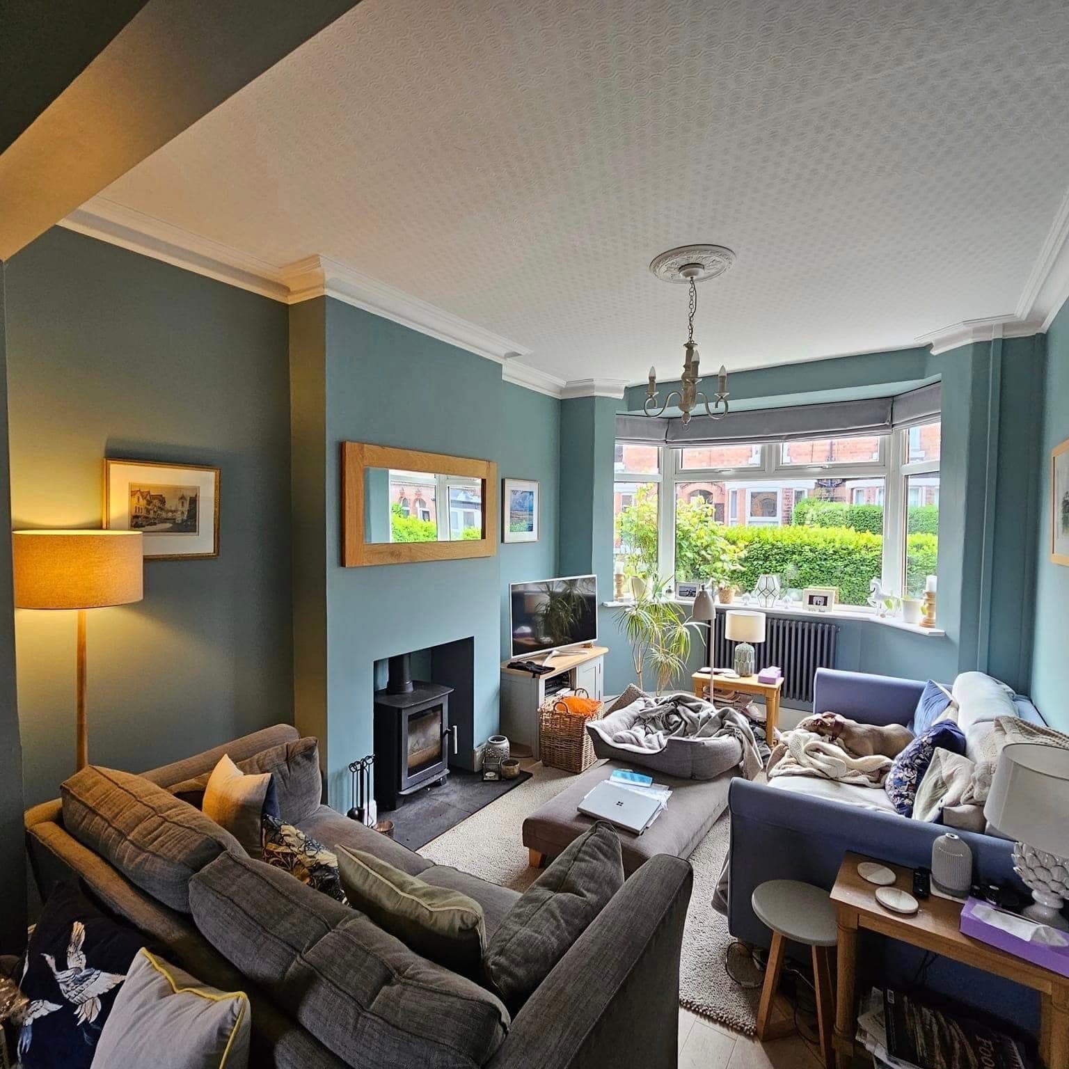 A cosy lounge in teal with a bay window, a wood burner and a framed mirror