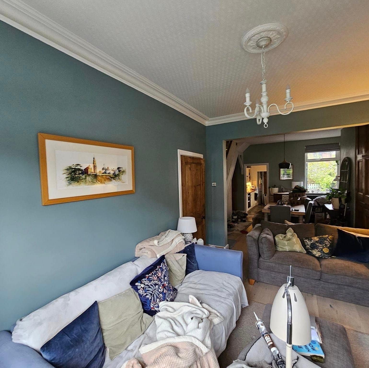 A lounge painted deep teal with a white ceiling rose, chandelier and coving, opening through to a dining room