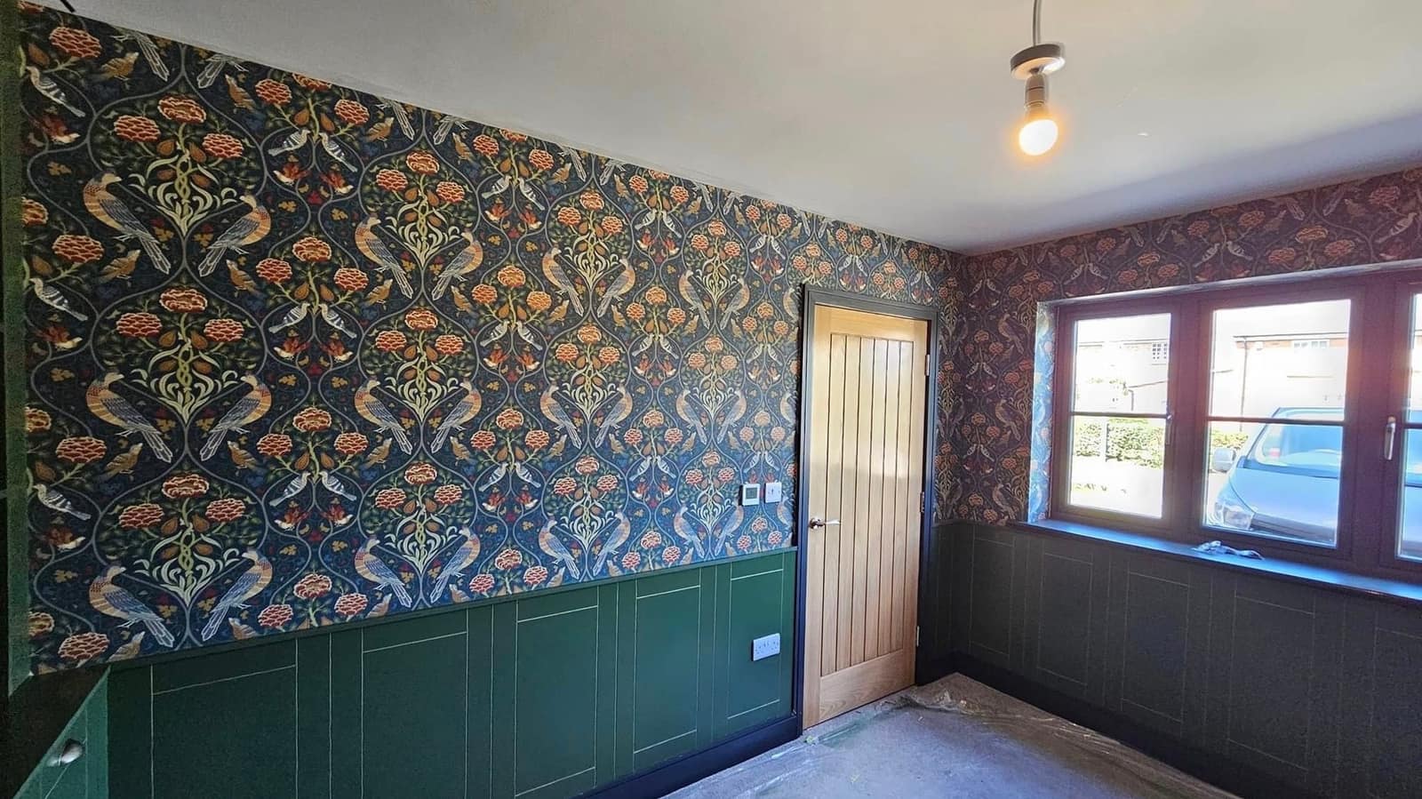 A room papered in a navy print of birds and foliage, above deep green panelling and beside an oak door