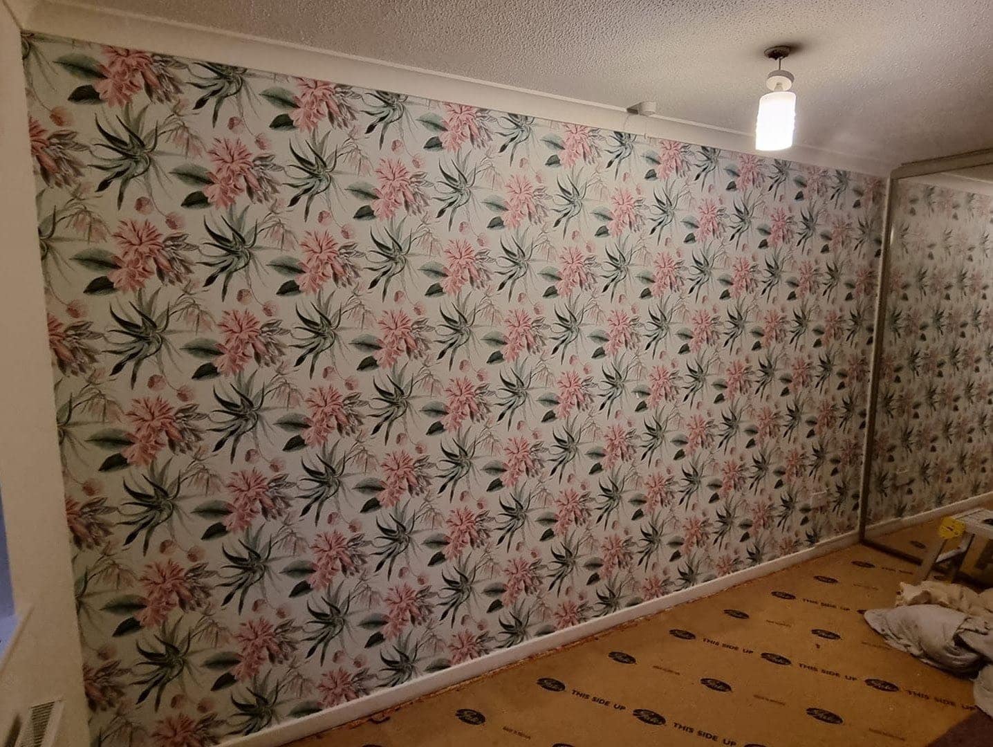 A long wall hung with a pink and green floral paper, the floor still protected