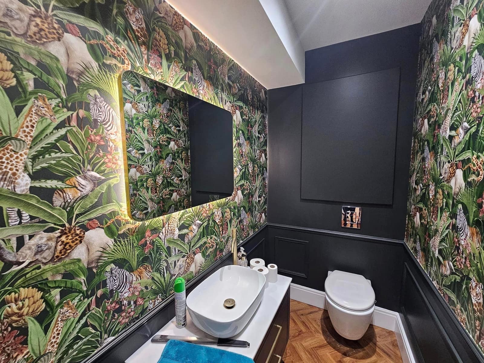 A cloakroom papered in a dense jungle print of giraffes, zebras and leaves, beside black-painted panelling and a backlit mirror
