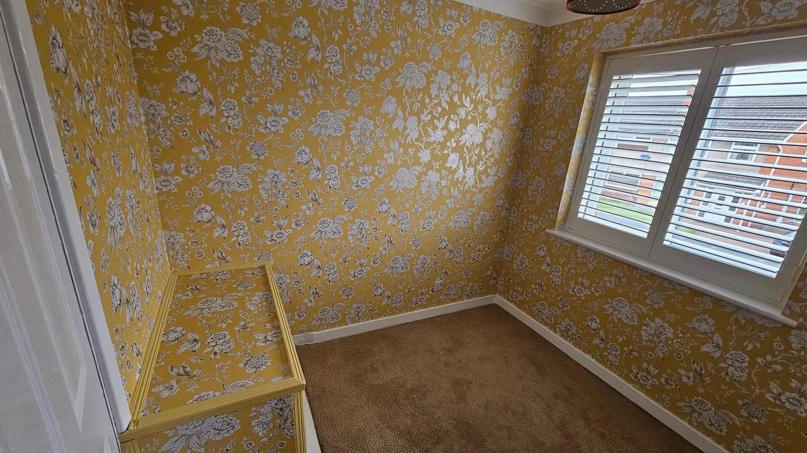 A bedroom papered in a mustard floral print on every wall, with white shutters at the window