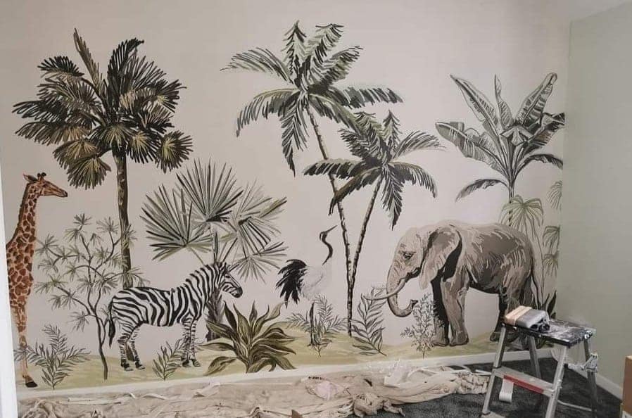 A botanical safari mural of palms, a giraffe, zebras and an elephant being hung, with the floor sheeted